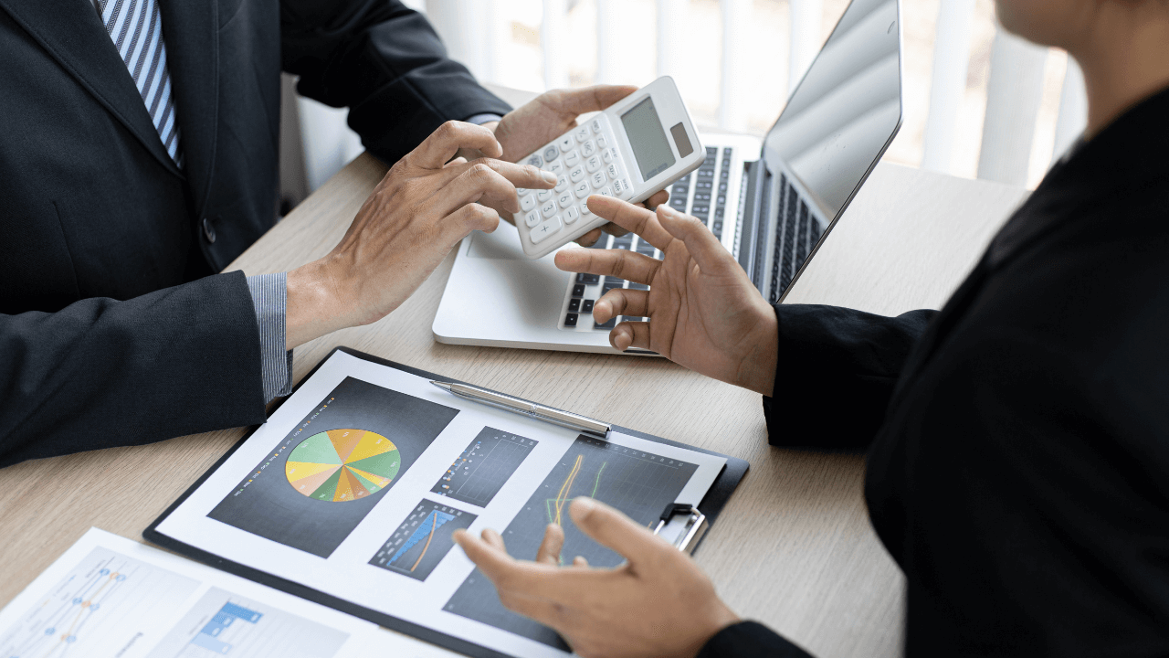 Business associates doing business over a desk of charts while holding a calculator | Scott Legal, P.C. Business associates doing business over a desk of charts while holding a calculator
