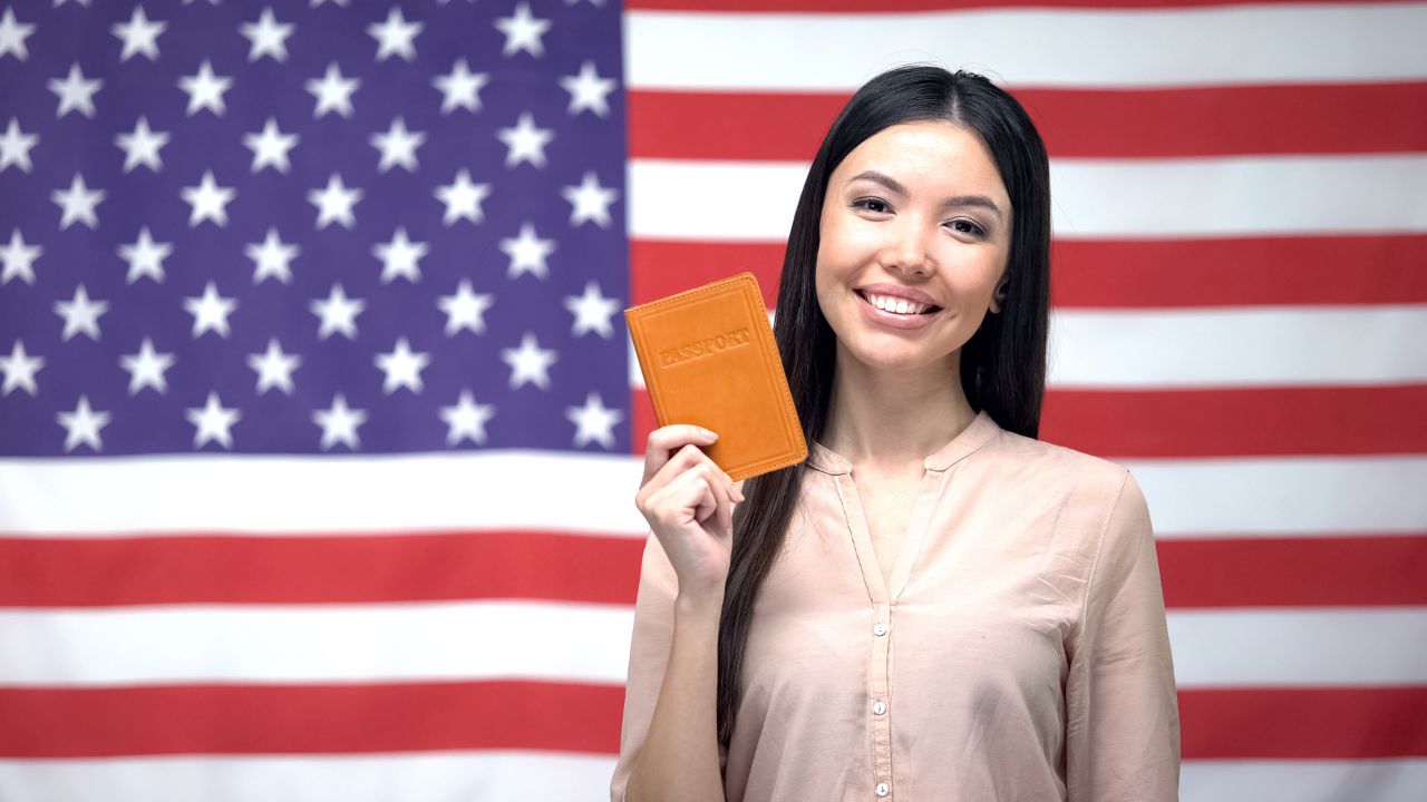 A woman smiling in front of a US flag while holding a passport | Scott Legal, P.C. A woman smiling in front of a US flag while holding a passport